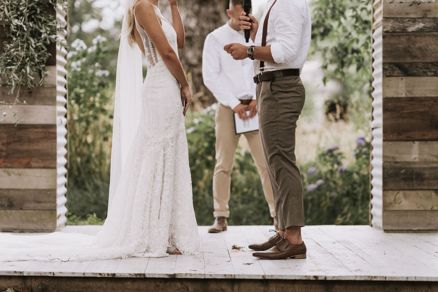 A wedding ceremony moment showing a white lace bridal gown and groom in khaki pants standing together at a rustic venue.