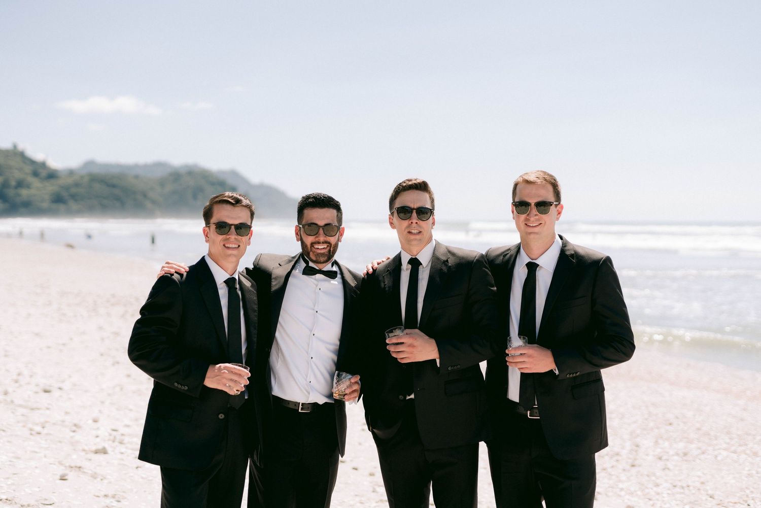 Four men in black suits and ties pose together on a sunny beach with ocean and mountains in the background.