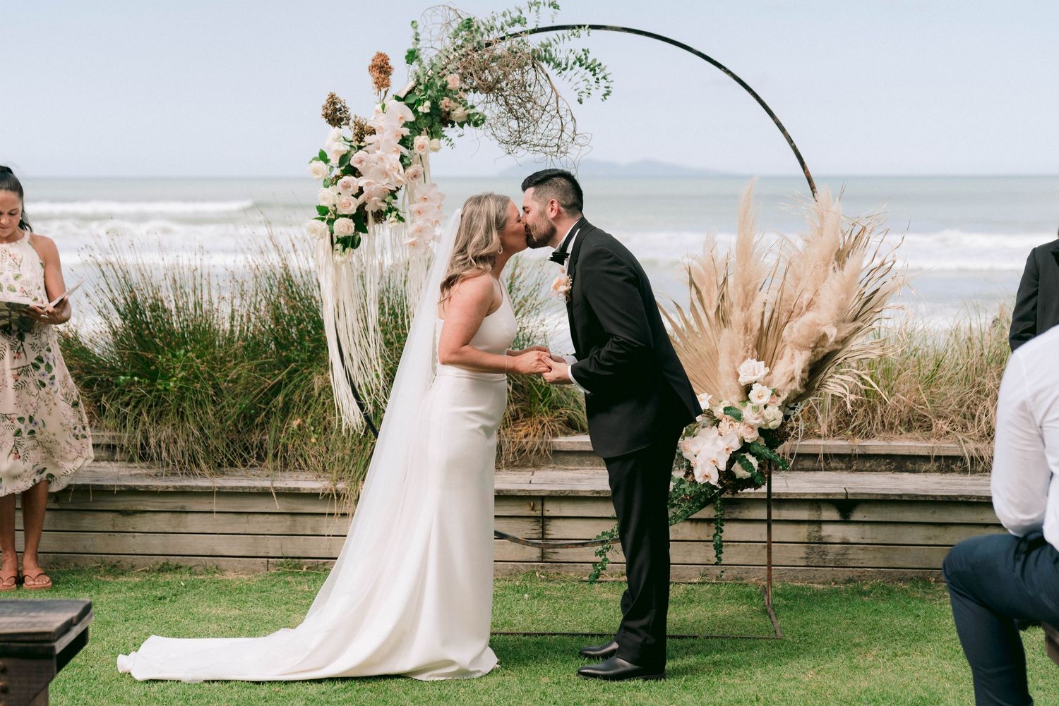 A seaside wedding ceremony under a floral arch with dried pampas grass and white roses on a grassy lawn by the beach.