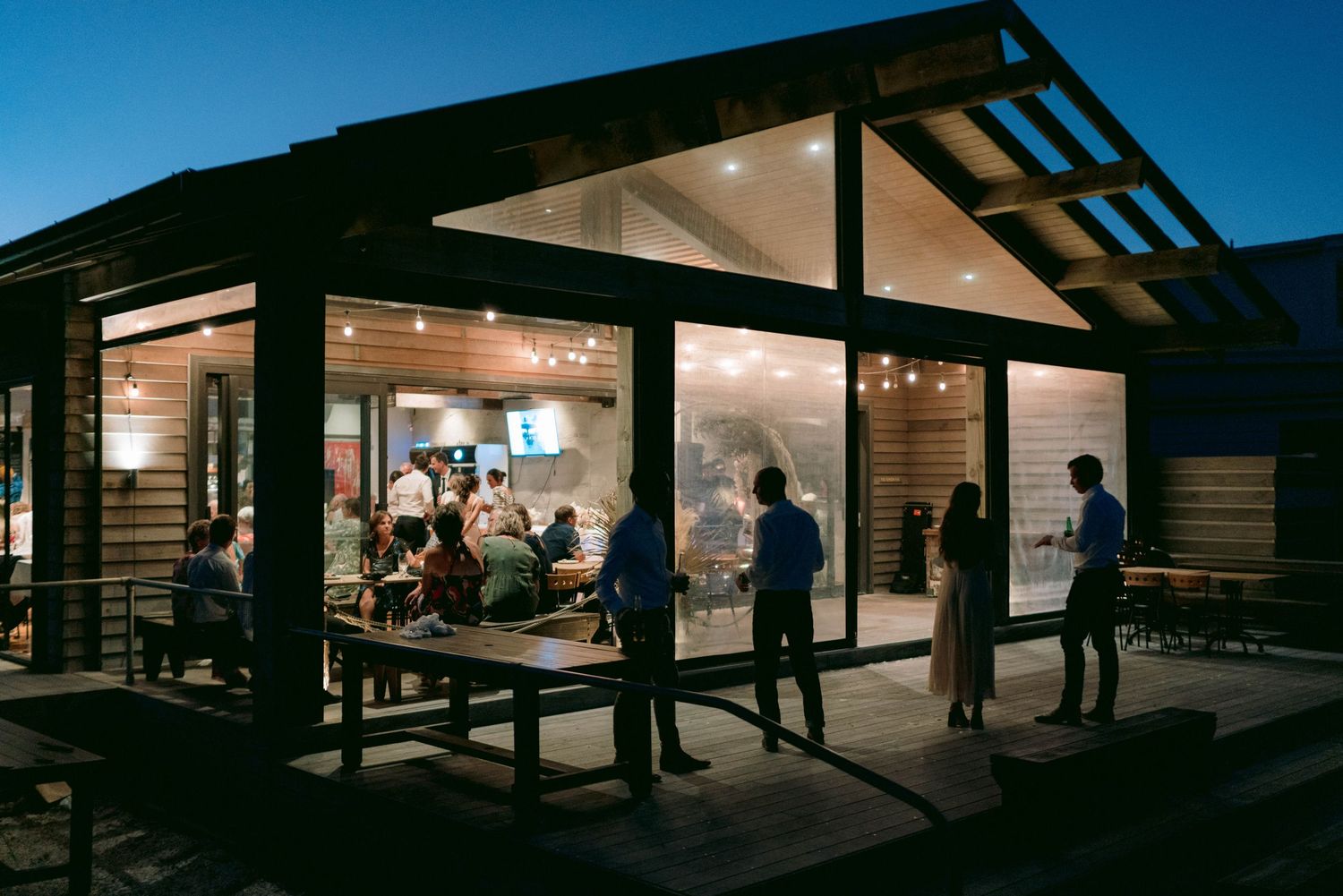 Evening wedding reception venue with guests mingling under string lights and dark wooden beams.