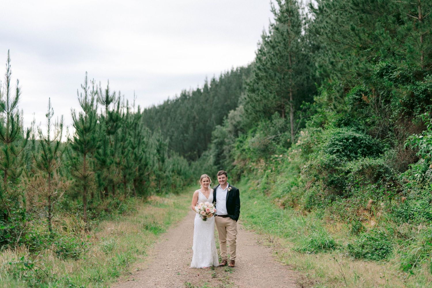 Newlyweds pose for a portrait on a mountain trail surrounded by tall pine trees.