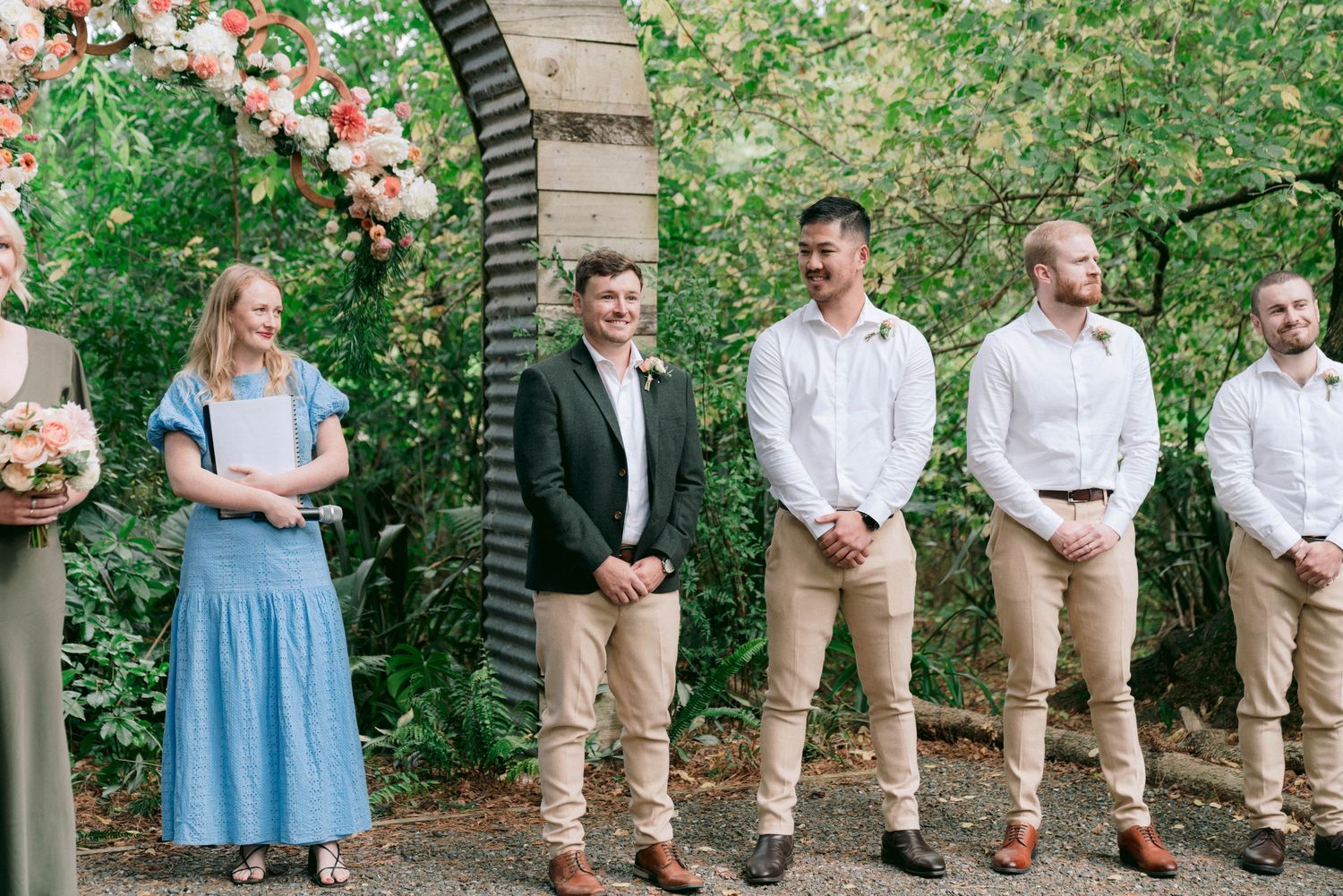 Wedding party lined up under floral arch wearing light blue and khaki attire.