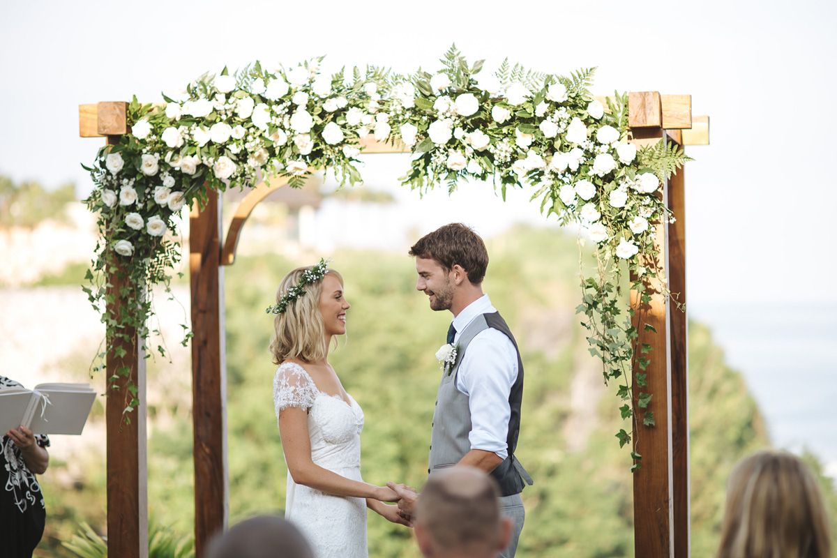 A romantic outdoor wedding ceremony under a wooden arch decorated with white floral arrangements and greenery.