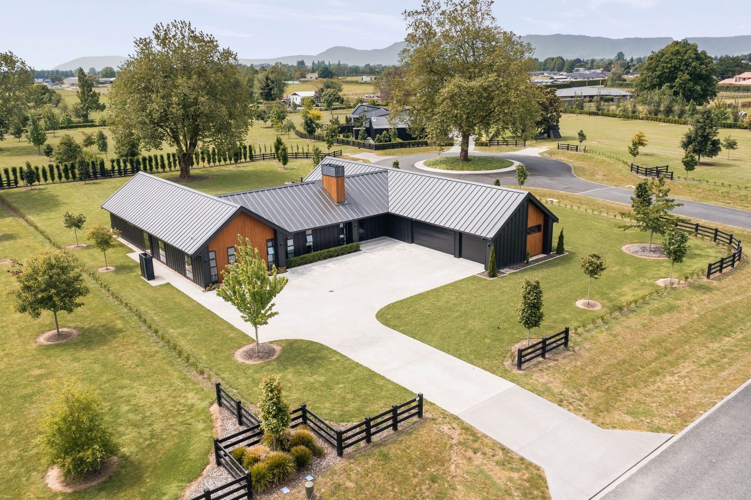 Drone shot of rural properties with long driveways, black barns and white fencing across rolling green landscape.