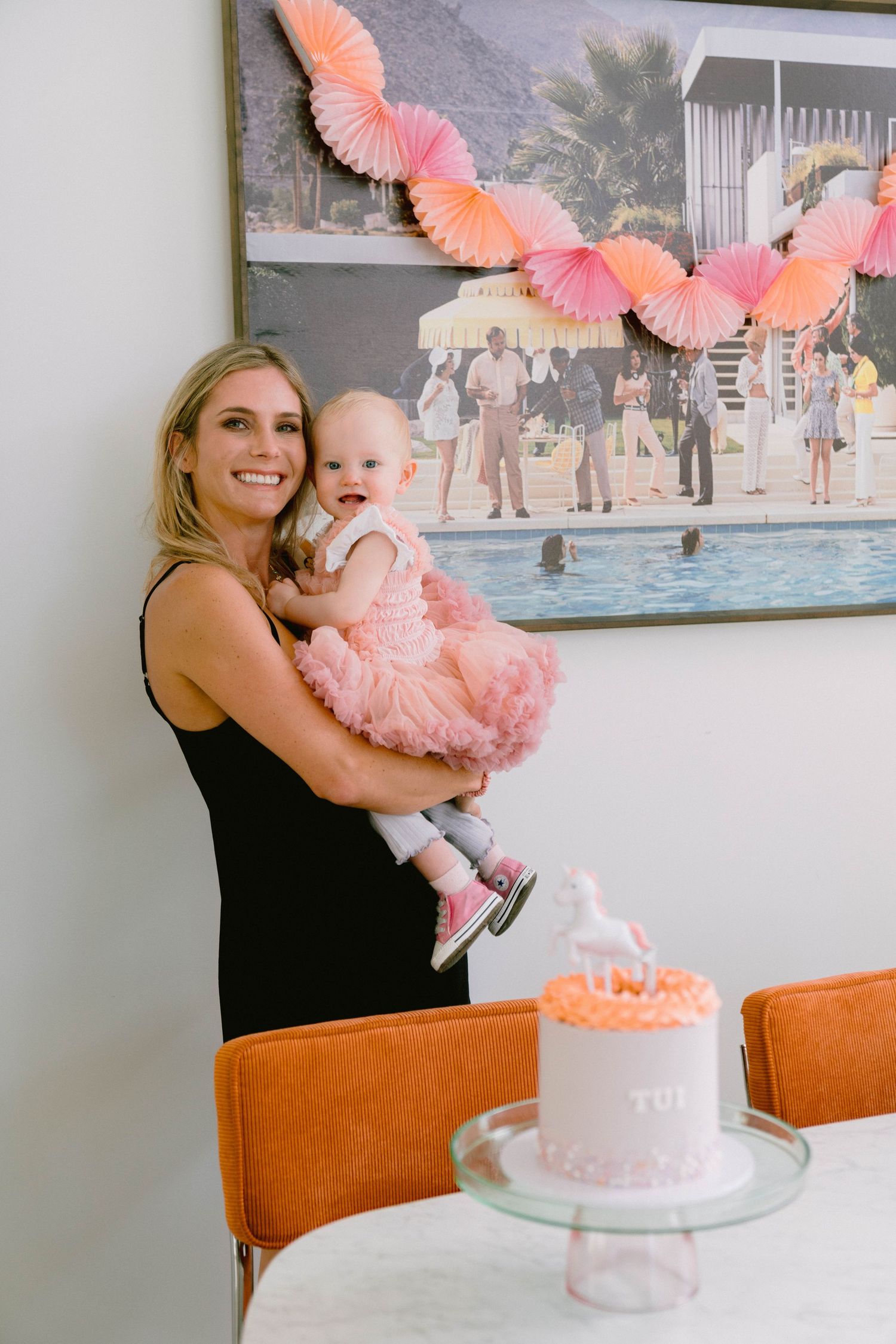 A celebratory scene with pink paper fan decorations and a small pink cake on a stand against a poolside backdrop artwork.