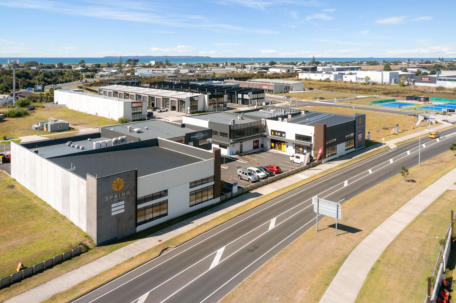 Modern commercial retail plaza with gray buildings and storefronts along a wide paved road viewed from above.