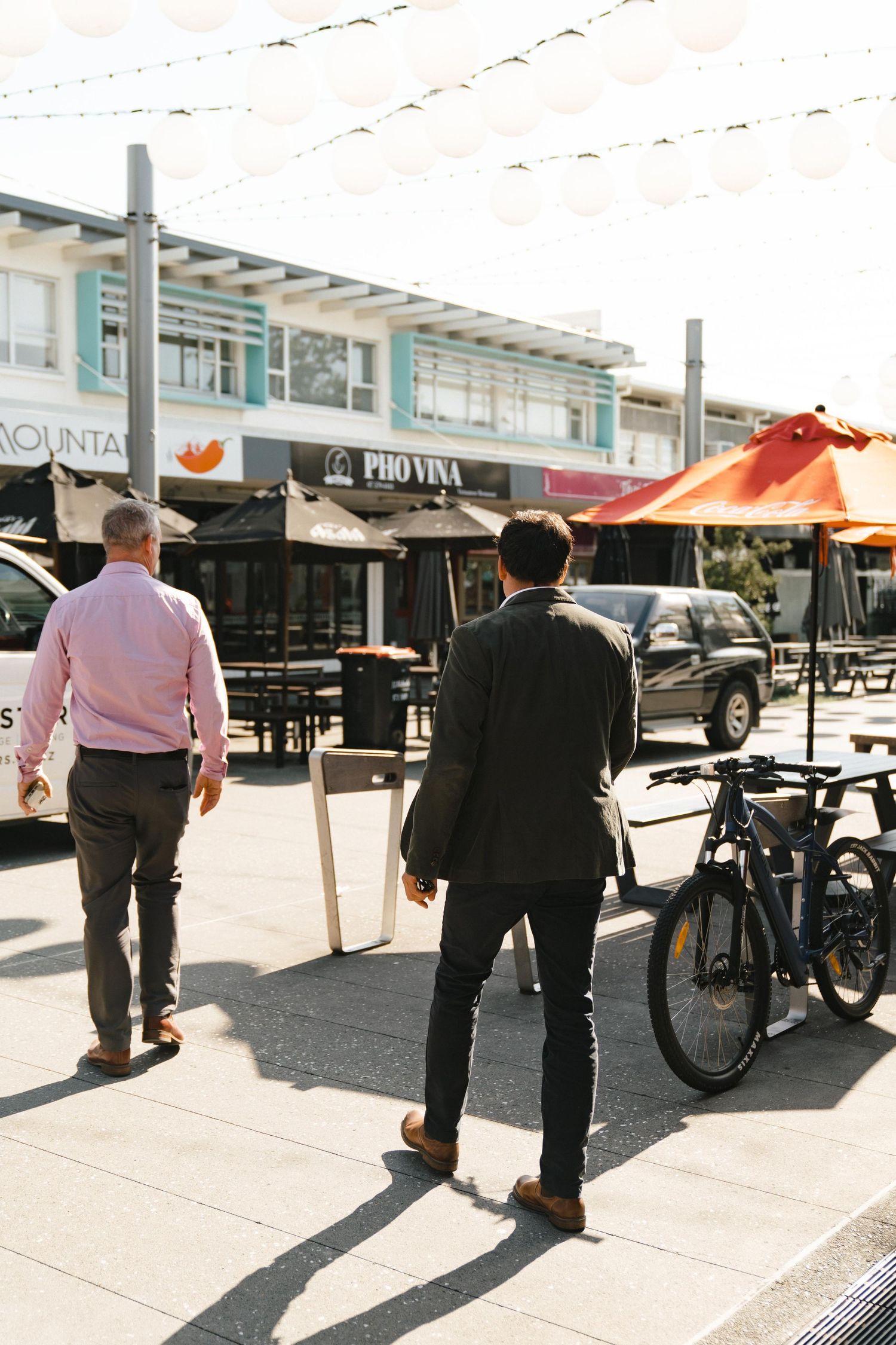 Two people walking past outdoor restaurant seating with umbrellas and a parked bicycle.
