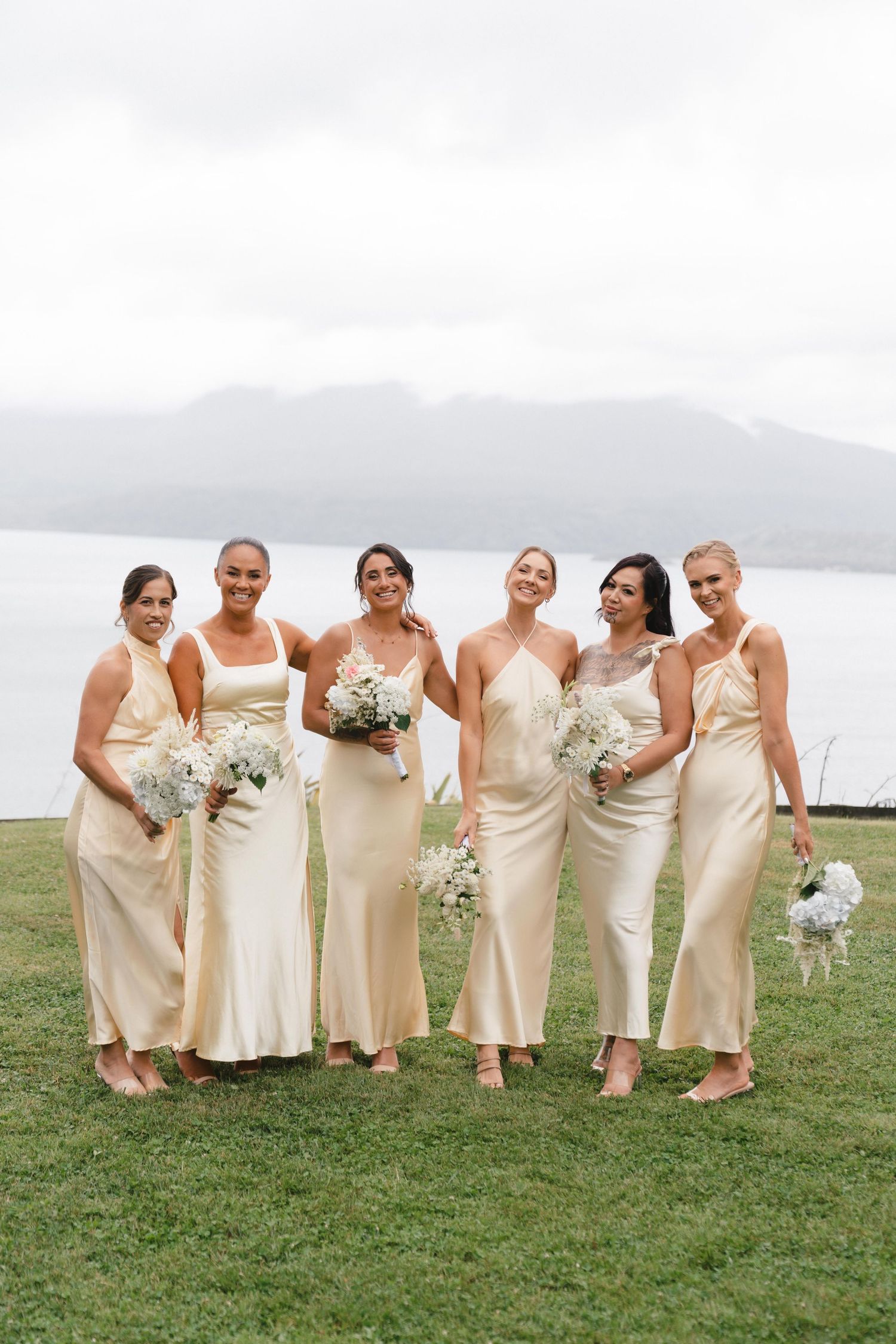 Bridesmaids in cream-colored dresses pose together on grass with mountain views and bouquets.