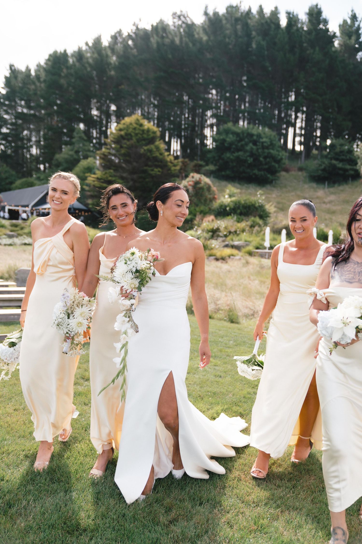 Bridesmaids in white dresses walking together across a grassy hill with pine trees in the background.