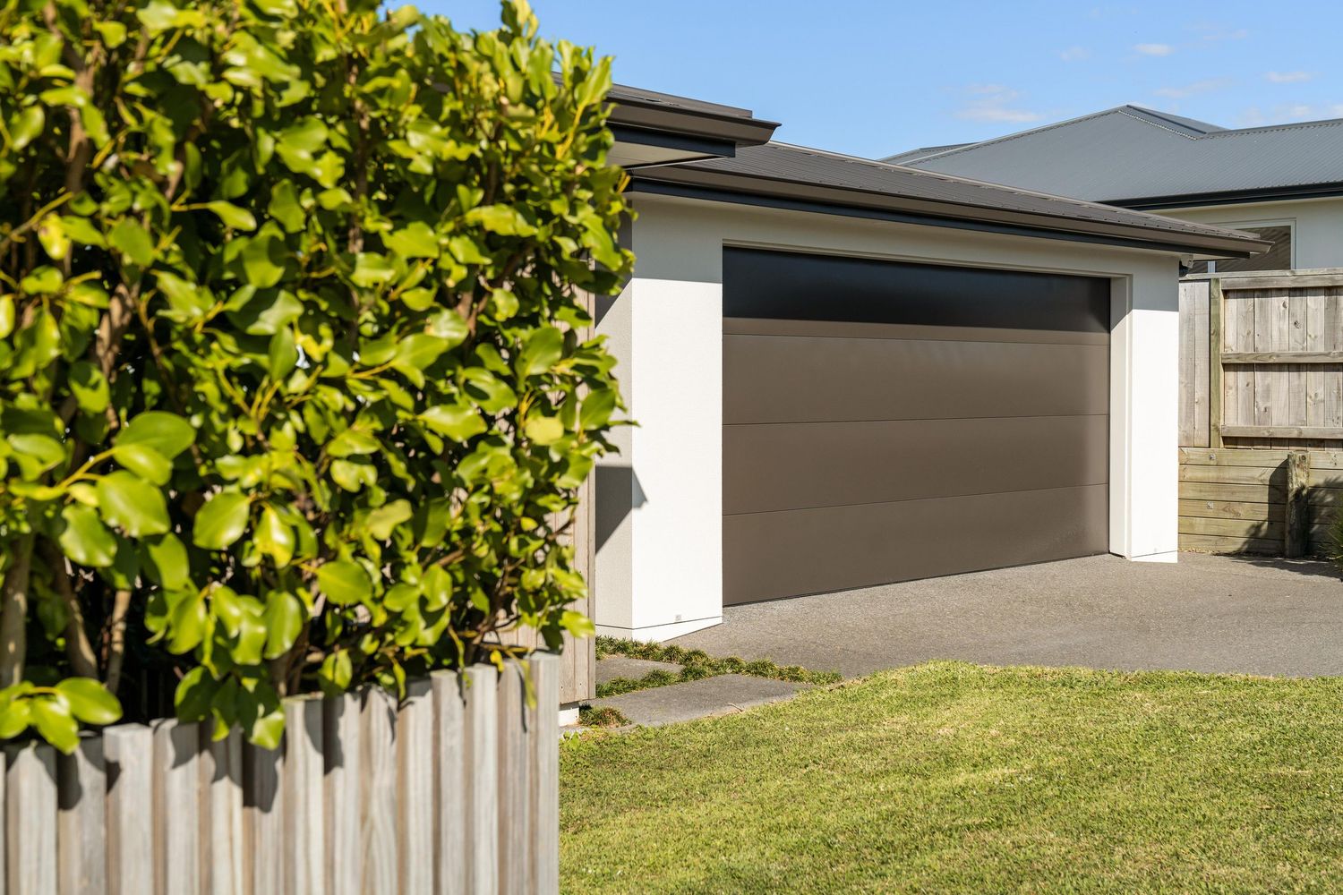 Modern brown garage door on residential home with green hedge and manicured lawn in foreground.