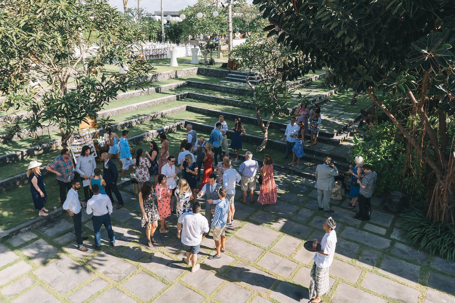Outdoor wedding guests gathering in a garden setting with trees and greenery.