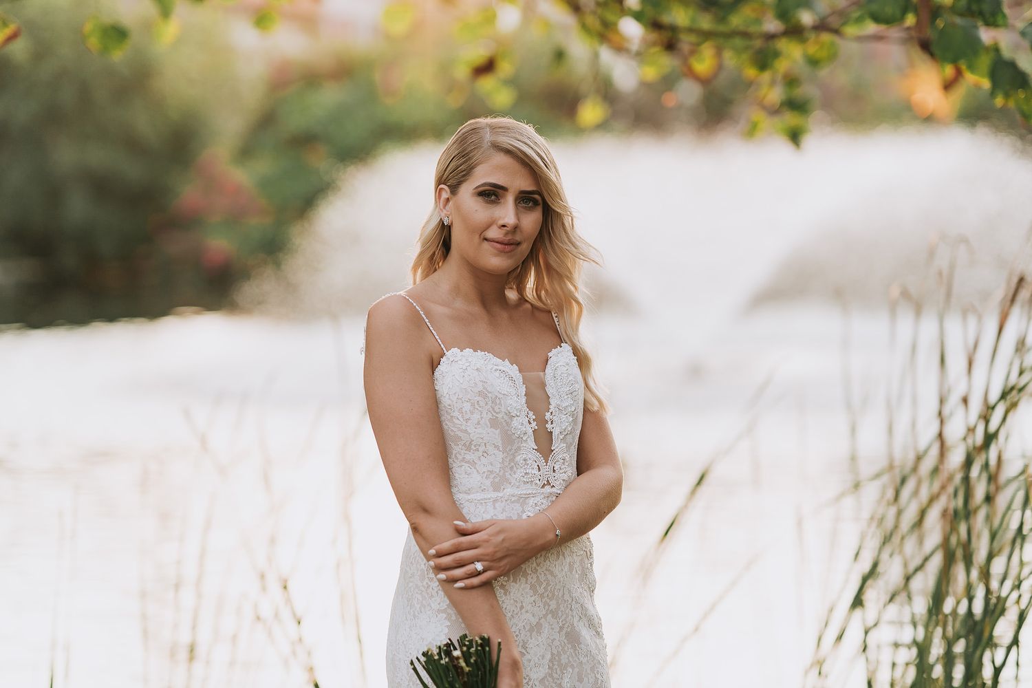 Elegant bride in lace wedding dress poses near lakeside reeds during golden hour outdoor photoshoot.