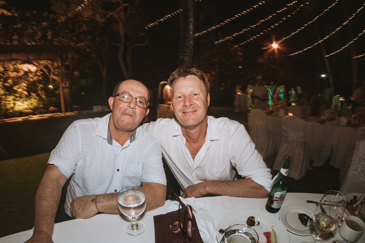 Two people seated at an outdoor dining table during an evening event with string lights overhead.