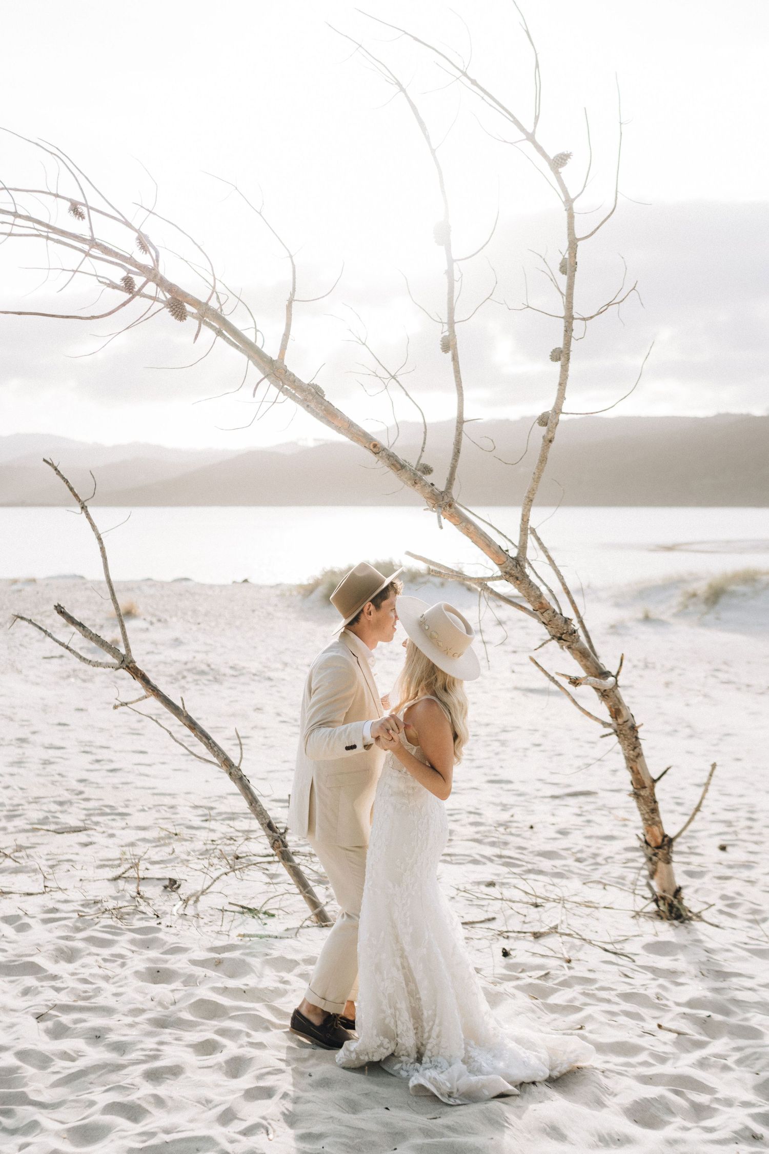 Couple in boho wedding attire embracing on sandy beach beside driftwood tree with mountain backdrop.