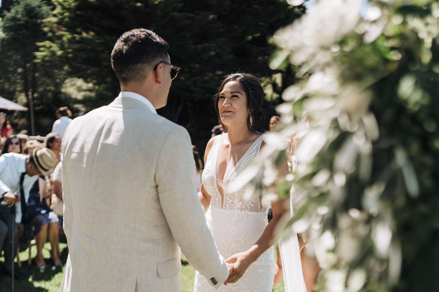 An intimate outdoor wedding ceremony takes place in dappled sunlight surrounded by greenery and guests.