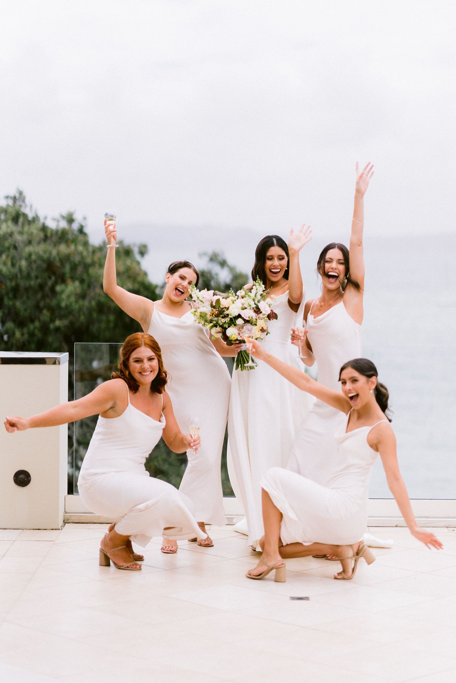 Bridesmaids in white dresses strike playful poses together on a bright outdoor terrace.