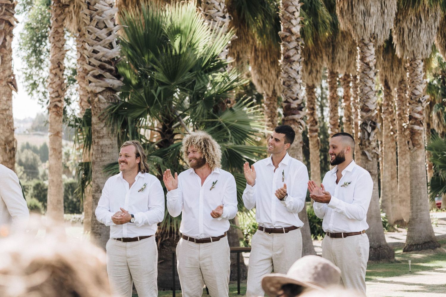 Groomsmen in white button-down shirts and slacks pose together between palm trees at an outdoor wedding venue.