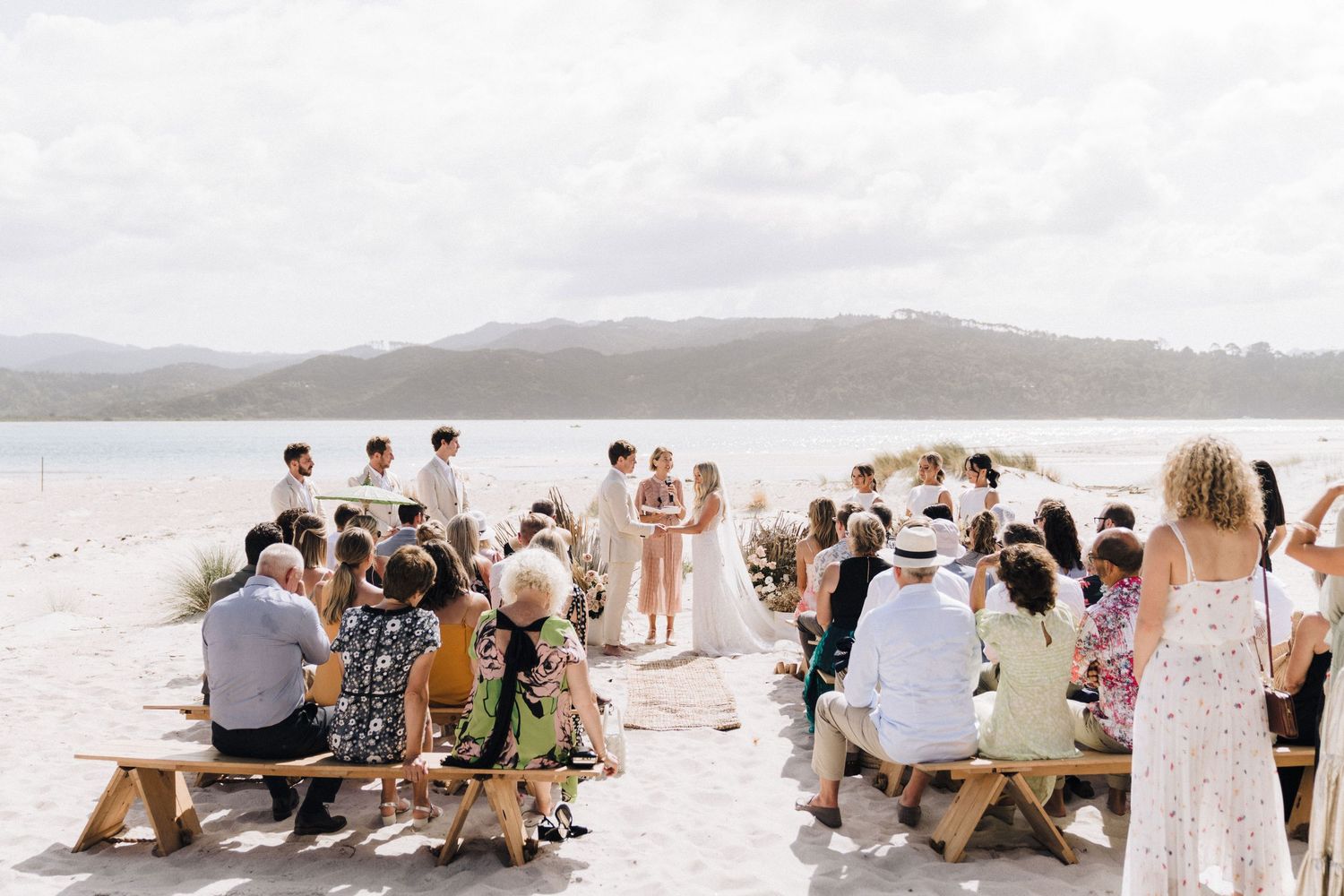 Outdoor wedding ceremony on a beach with guests seated on wooden benches overlooking a bay.