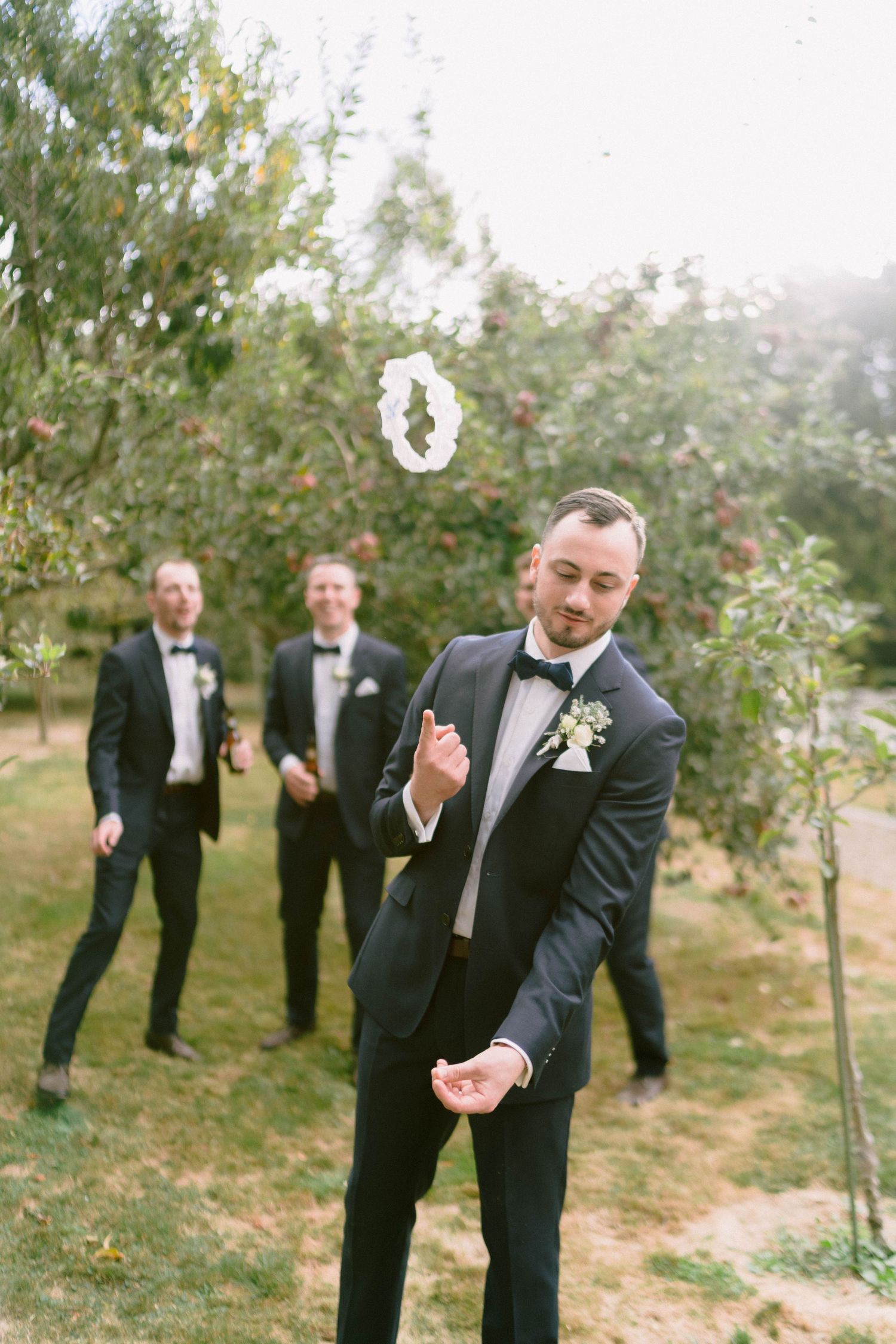 Groomsmen in black tuxedos pose playfully in an orchard setting during a wedding.