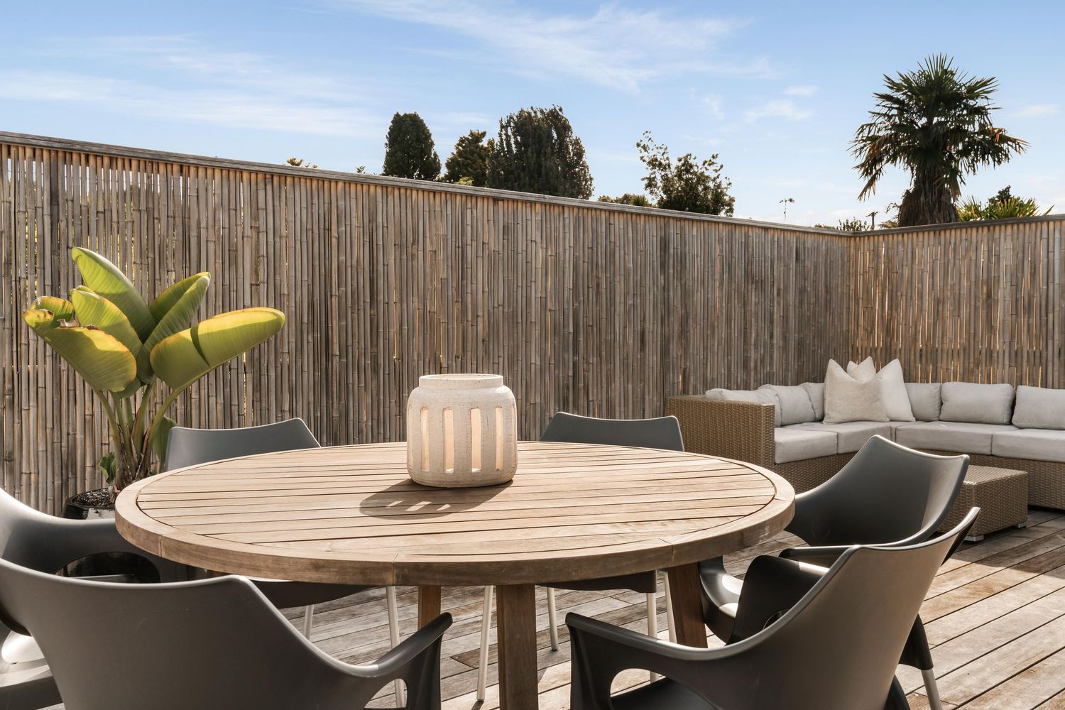 Round wooden outdoor dining table with grey chairs on a deck with bamboo fencing.