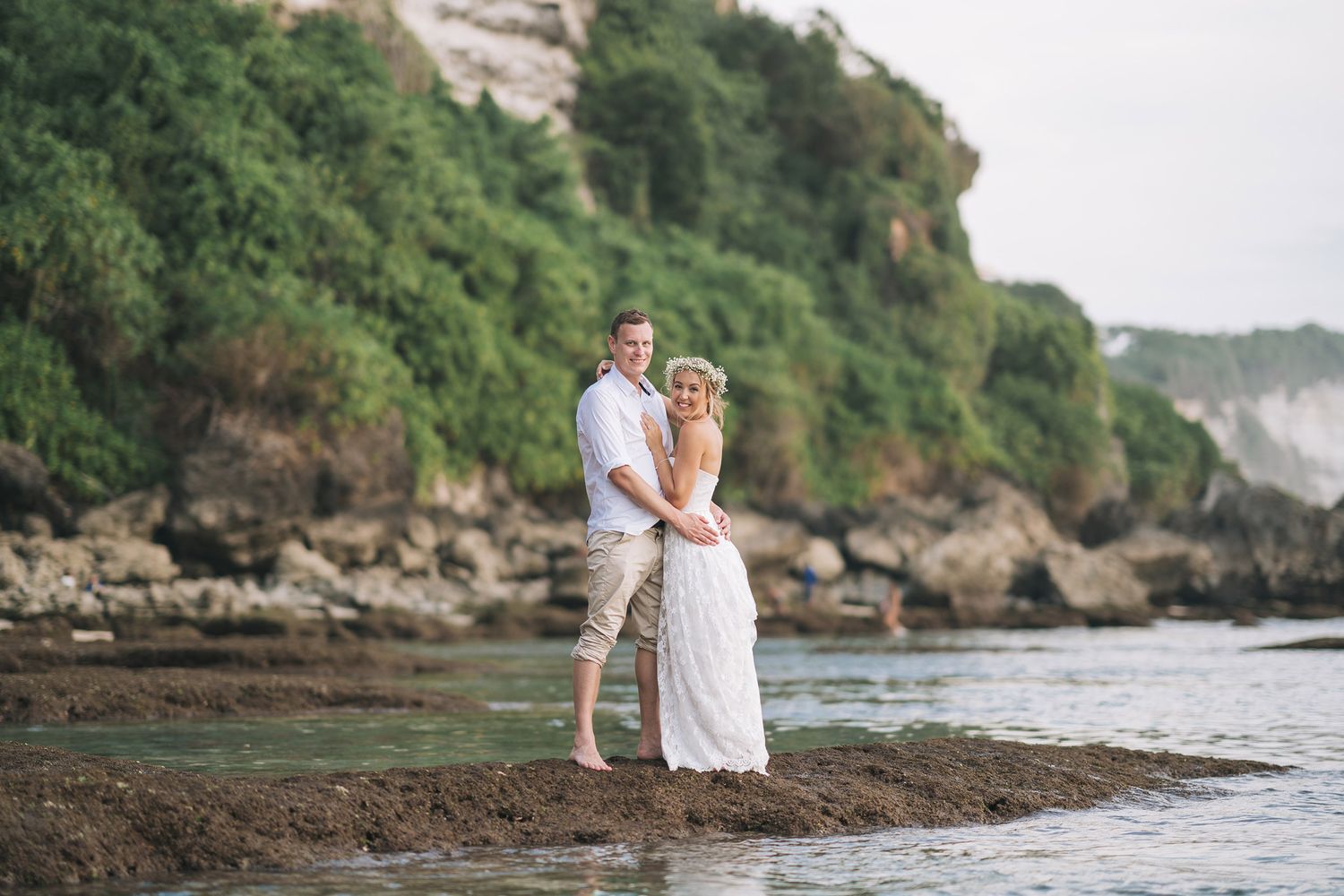 A newlywed couple poses on a beach with rocky cliffs and green vegetation in the background.