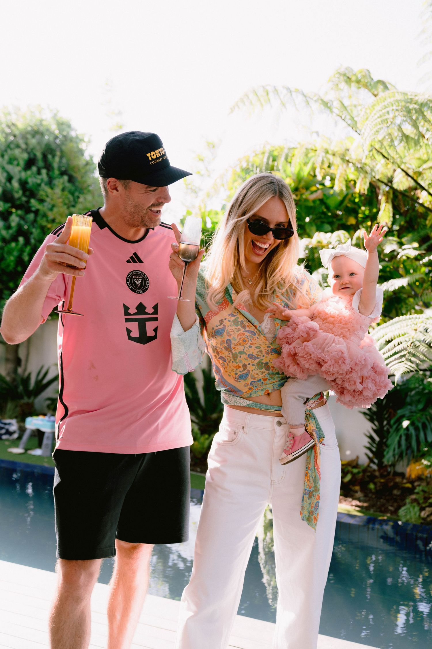 Friends enjoying drinks by the pool on a sunny day surrounded by tropical landscaping.