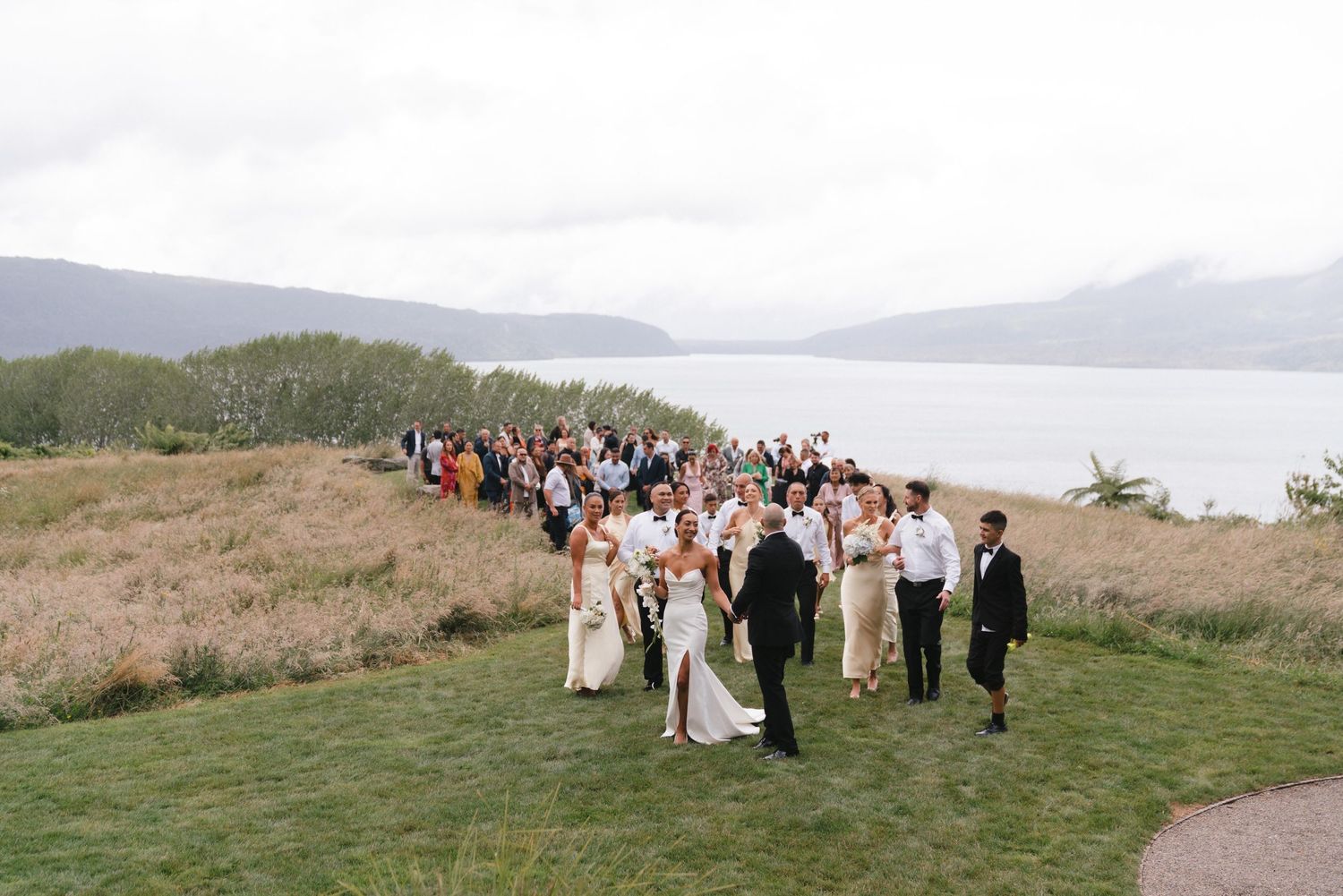 Wedding party and guests gather on a coastal hillside with water views in background.