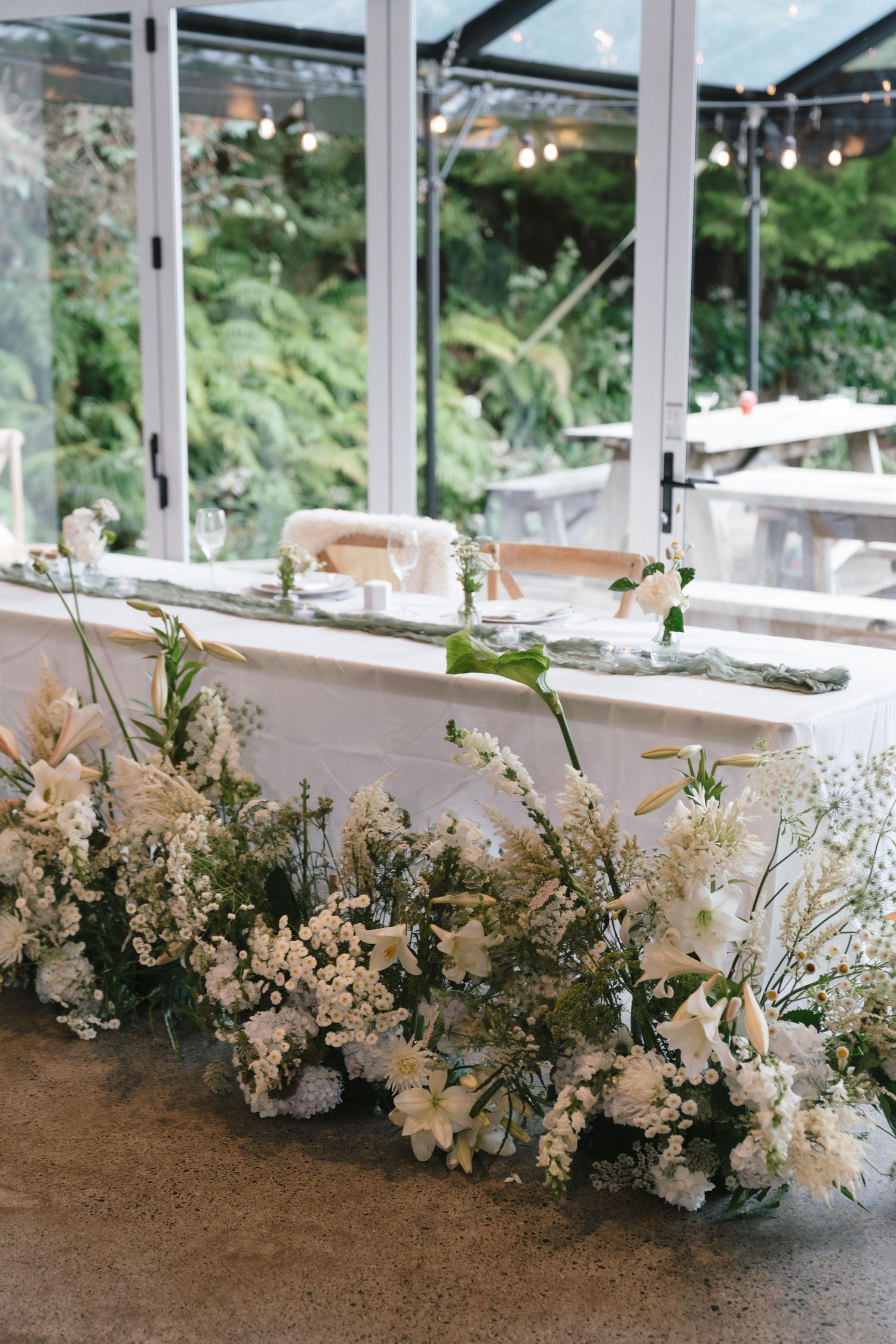 Elegant white floral arrangements line a glass-enclosed reception venue table.