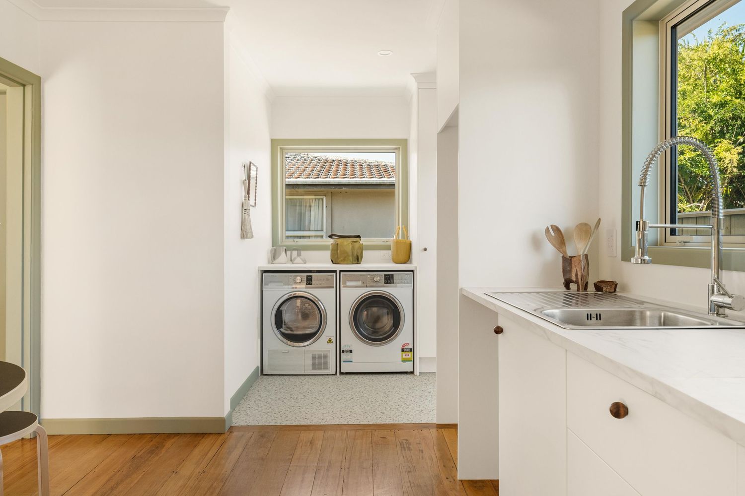 Modern laundry nook with stacked washer and dryer built into white wall, next to bright kitchen with hardwood floors.