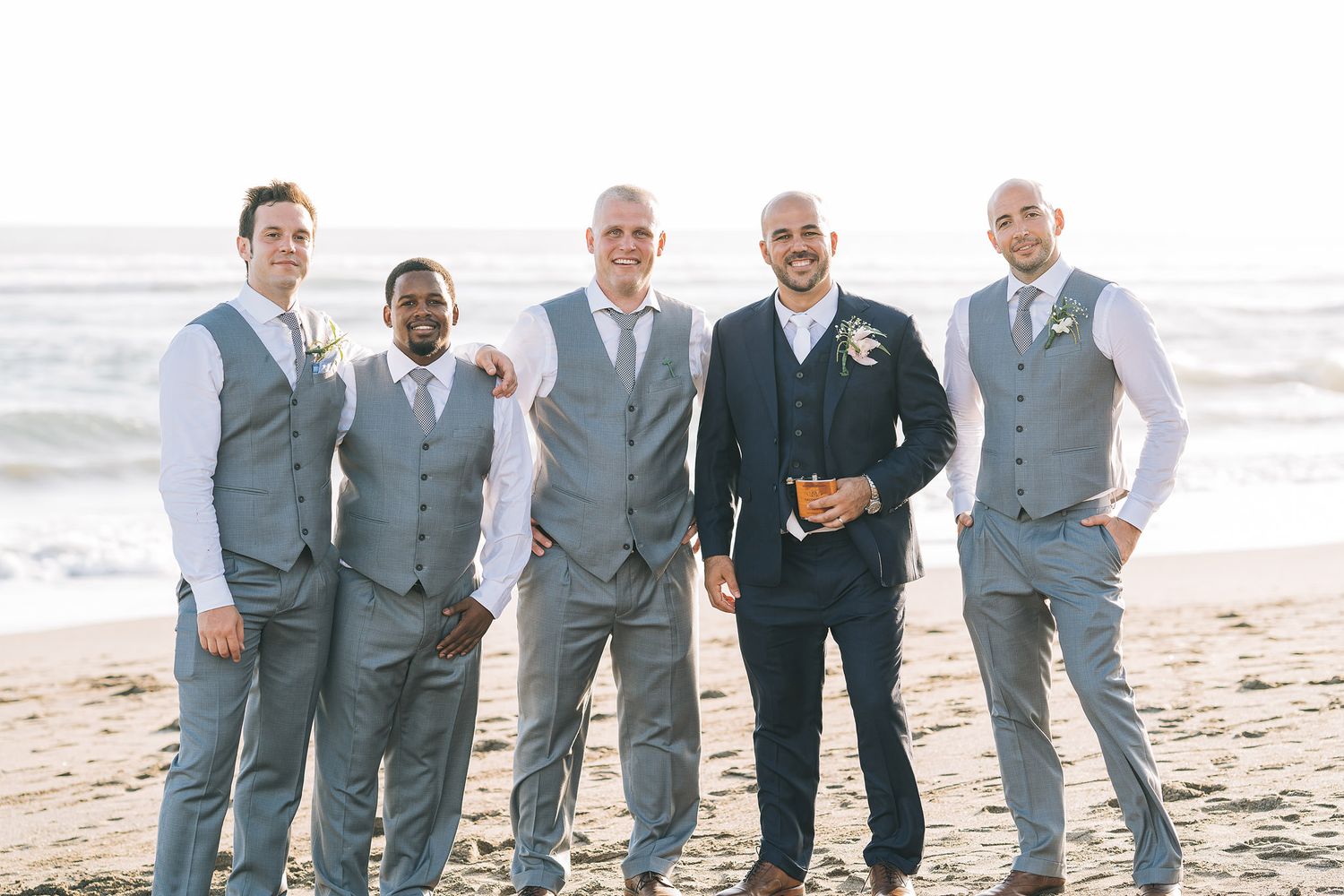 Groomsmen in matching grey suits pose together on a beach.