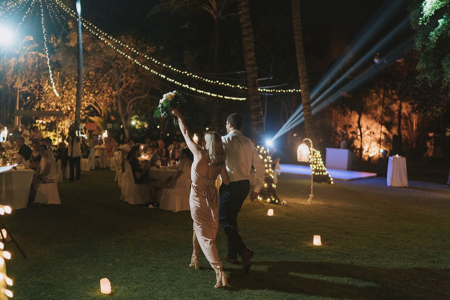 Evening outdoor wedding reception with string lights and guests dancing under palm trees.