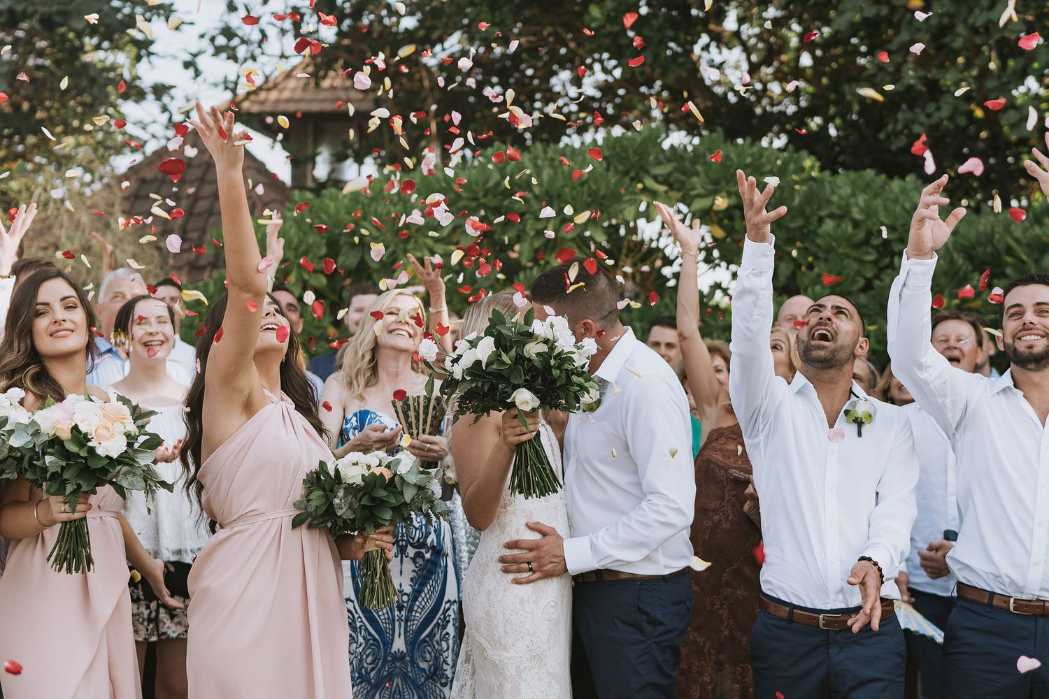 Wedding party celebrates with confetti toss under trees during outdoor reception.