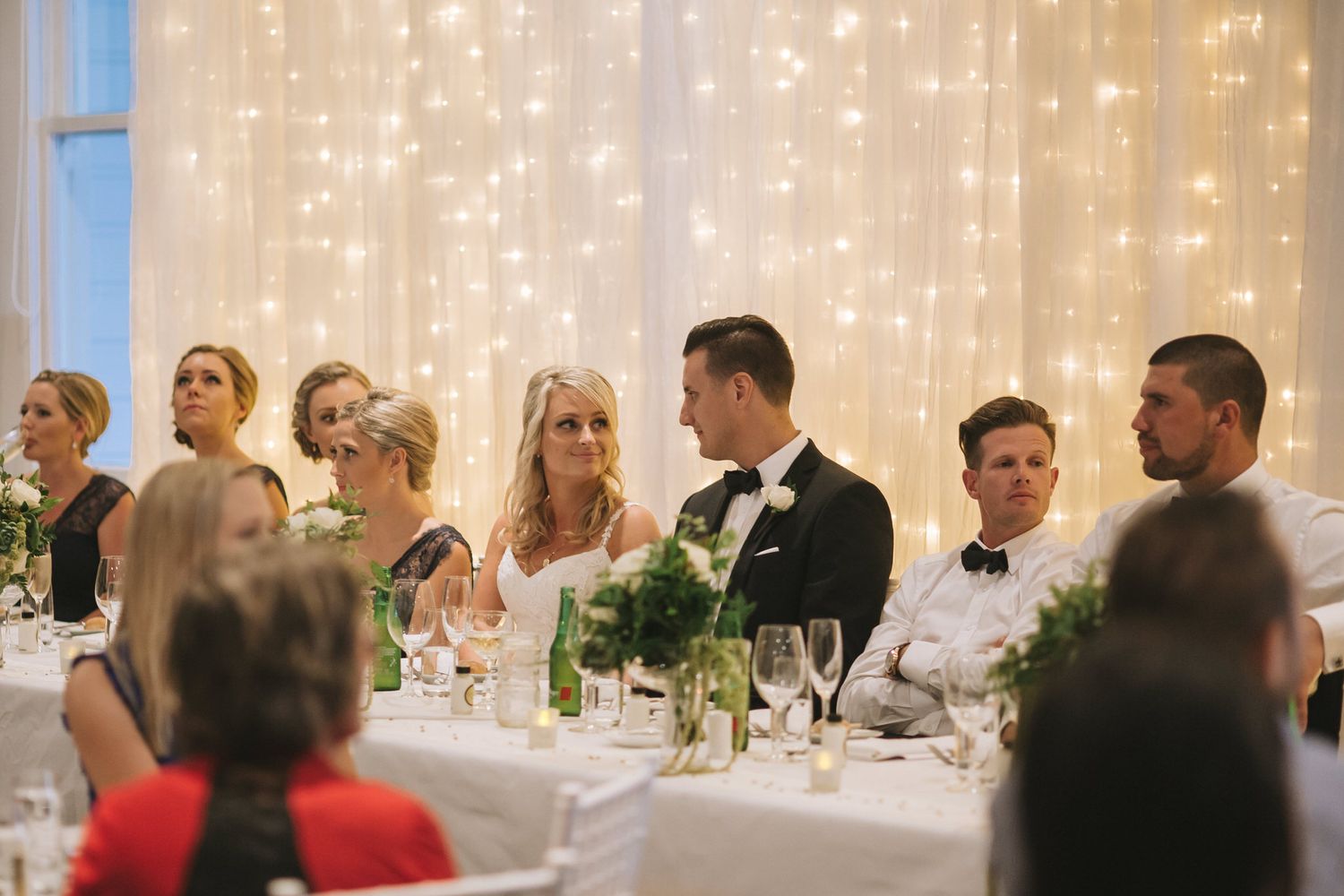 Wedding guests seated at elegant tables with white decor and twinkle lights creating a romantic reception atmosphere.
