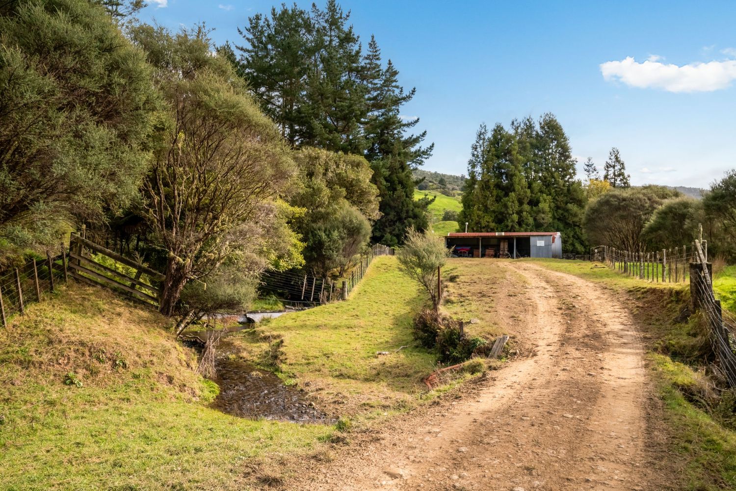 A winding dirt road leads through green countryside to a small shed or shelter in the distance on a sunny day.