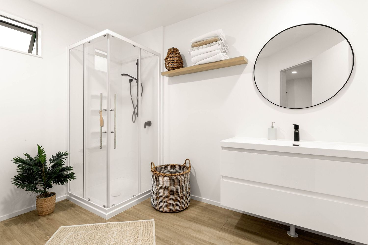 Modern white bathroom interior with glass shower, floating vanity, round mirror and wooden accents on herringbone flooring.
