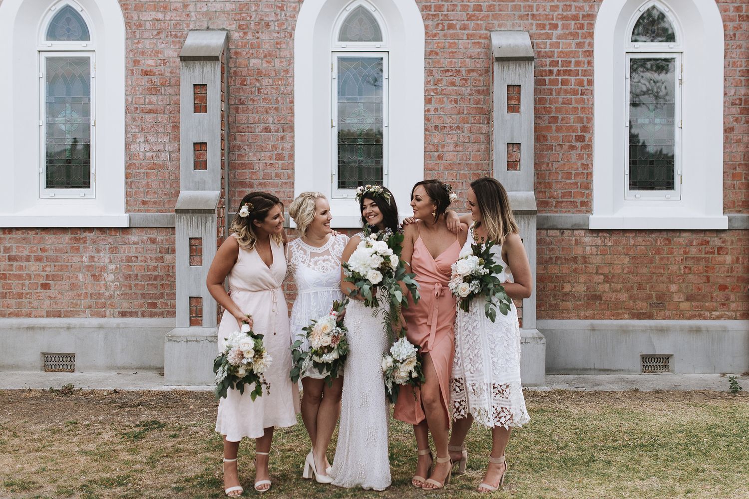 Five women in elegant white and blush dresses pose together outside a brick building with arched windows.