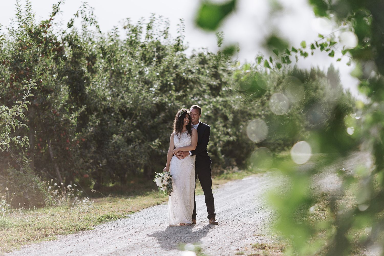A couple in wedding attire embraces on a sunlit gravel path surrounded by lush greenery and sparkling bokeh effects.