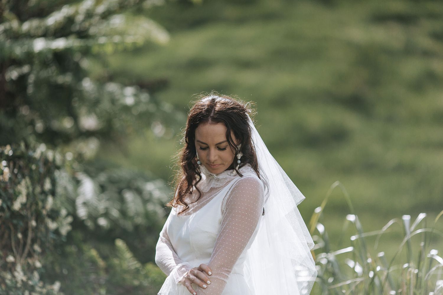 A bride in a white lace dress stands thoughtfully in a meadow surrounded by wildflowers and soft green foliage.
