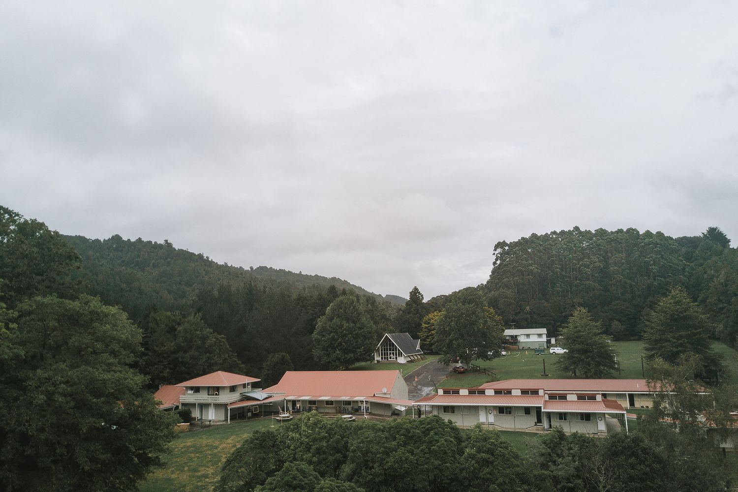 Rural motel or resort buildings with terracotta roofs nestled among wooded hills under an overcast sky.