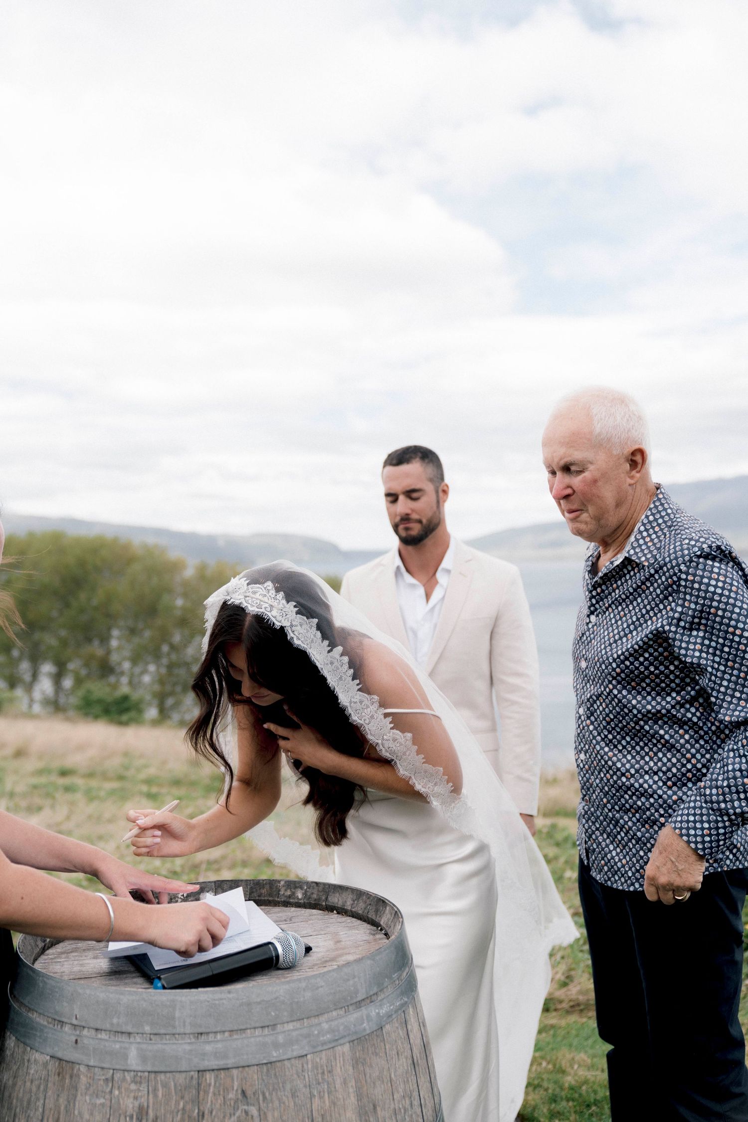 Wedding ceremony participants gather around a rustic wine barrel under cloudy skies.