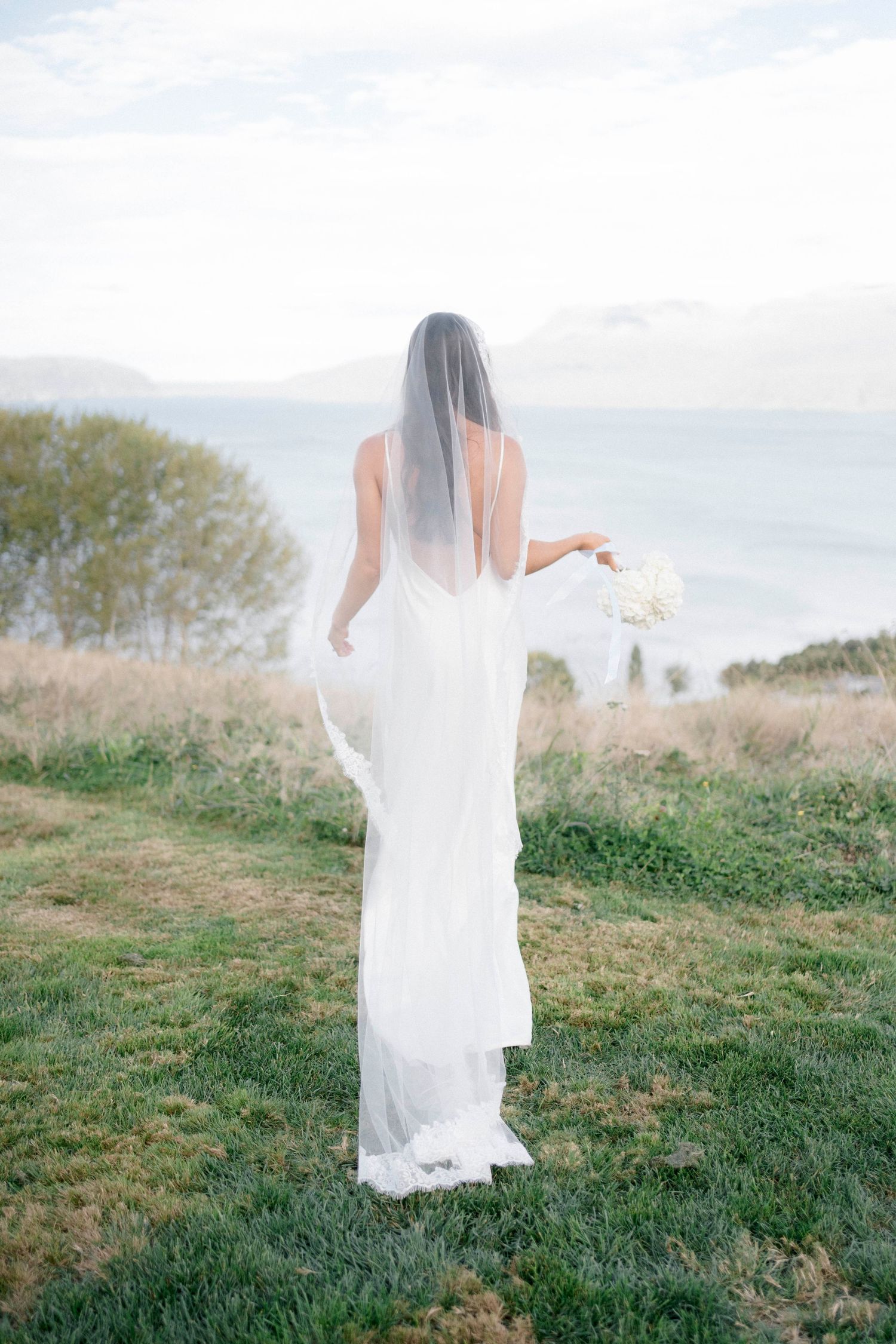 A bride in flowing white dress walking through a grassy field with veil trailing behind.