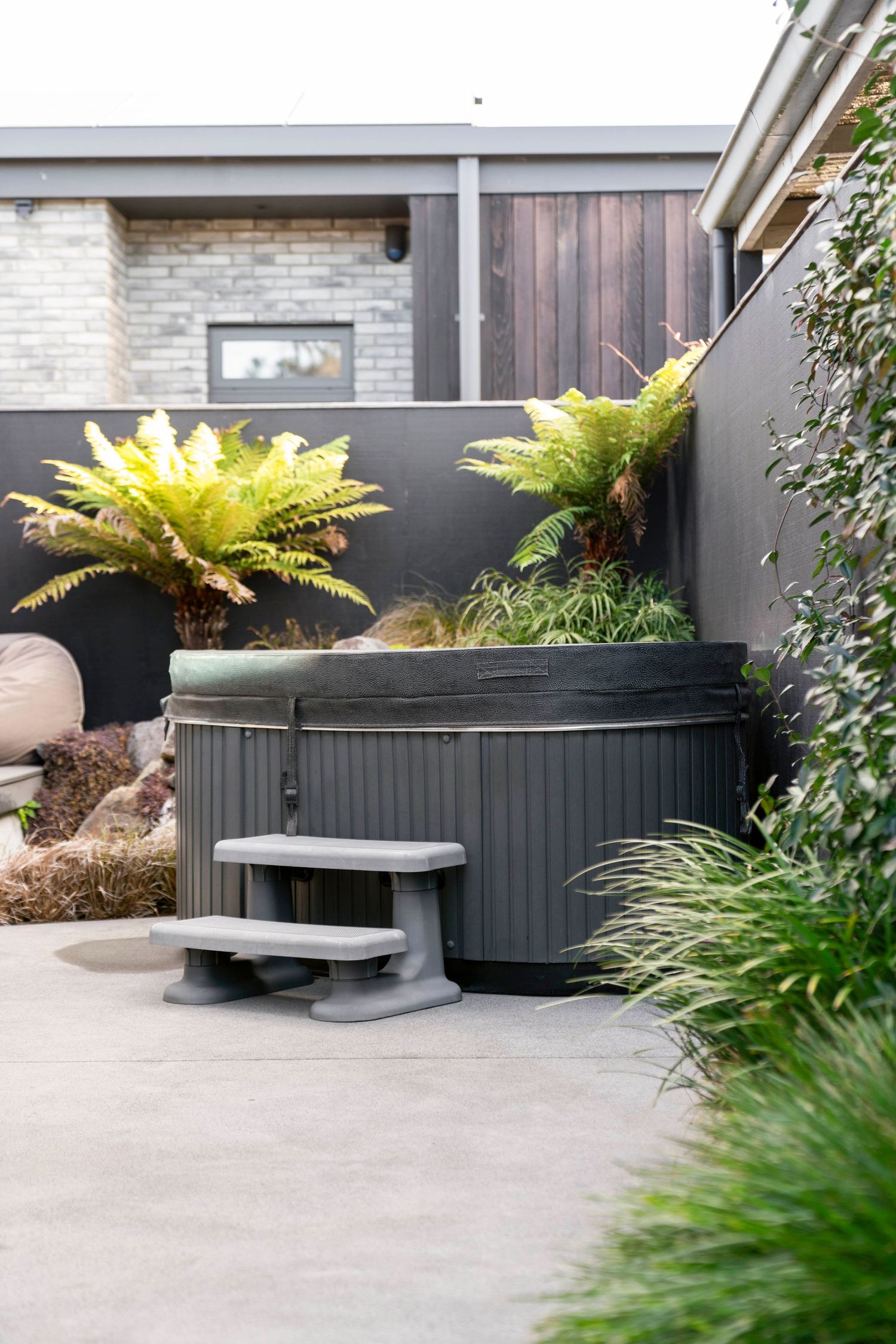 Modern outdoor hot tub with charcoal grey panels surrounded by ferns and tropical plants on a concrete patio.