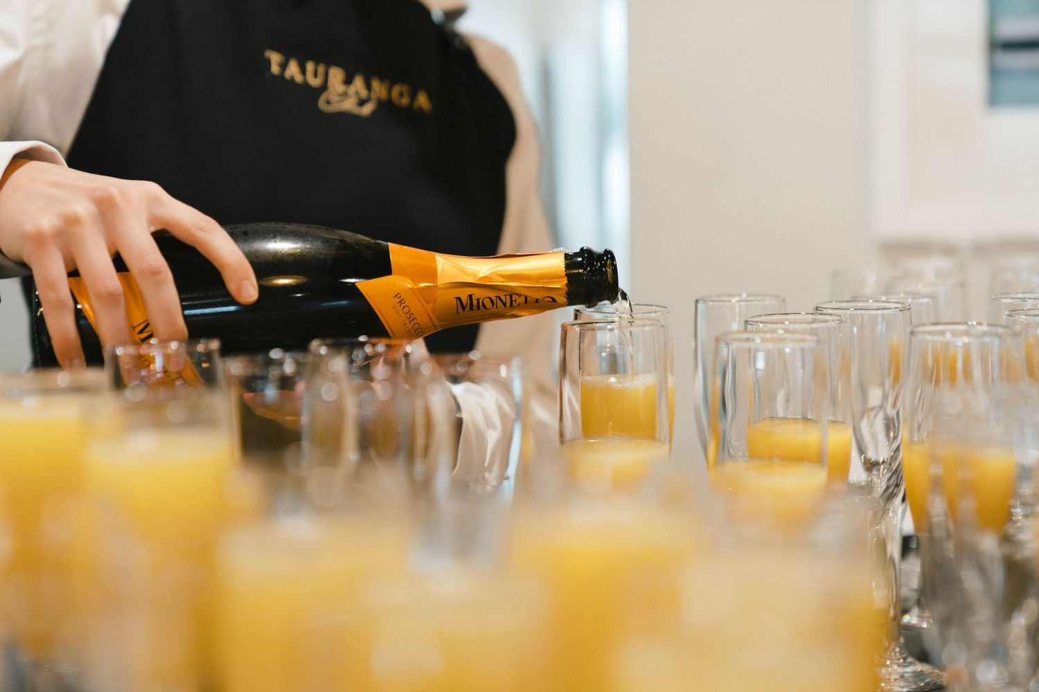 Orange juice is being poured into multiple champagne glasses lined up on a surface during cocktail service preparation.
