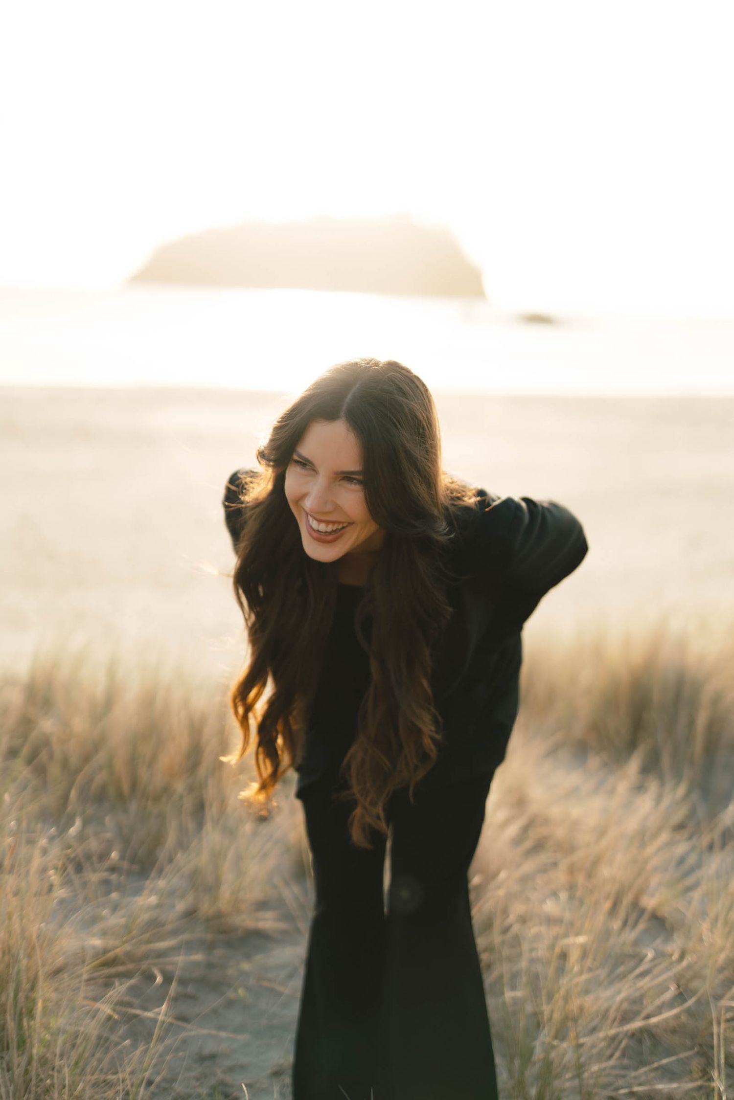 A person in a black sweater smiles joyfully while standing in a golden sun-lit field of tall grass.