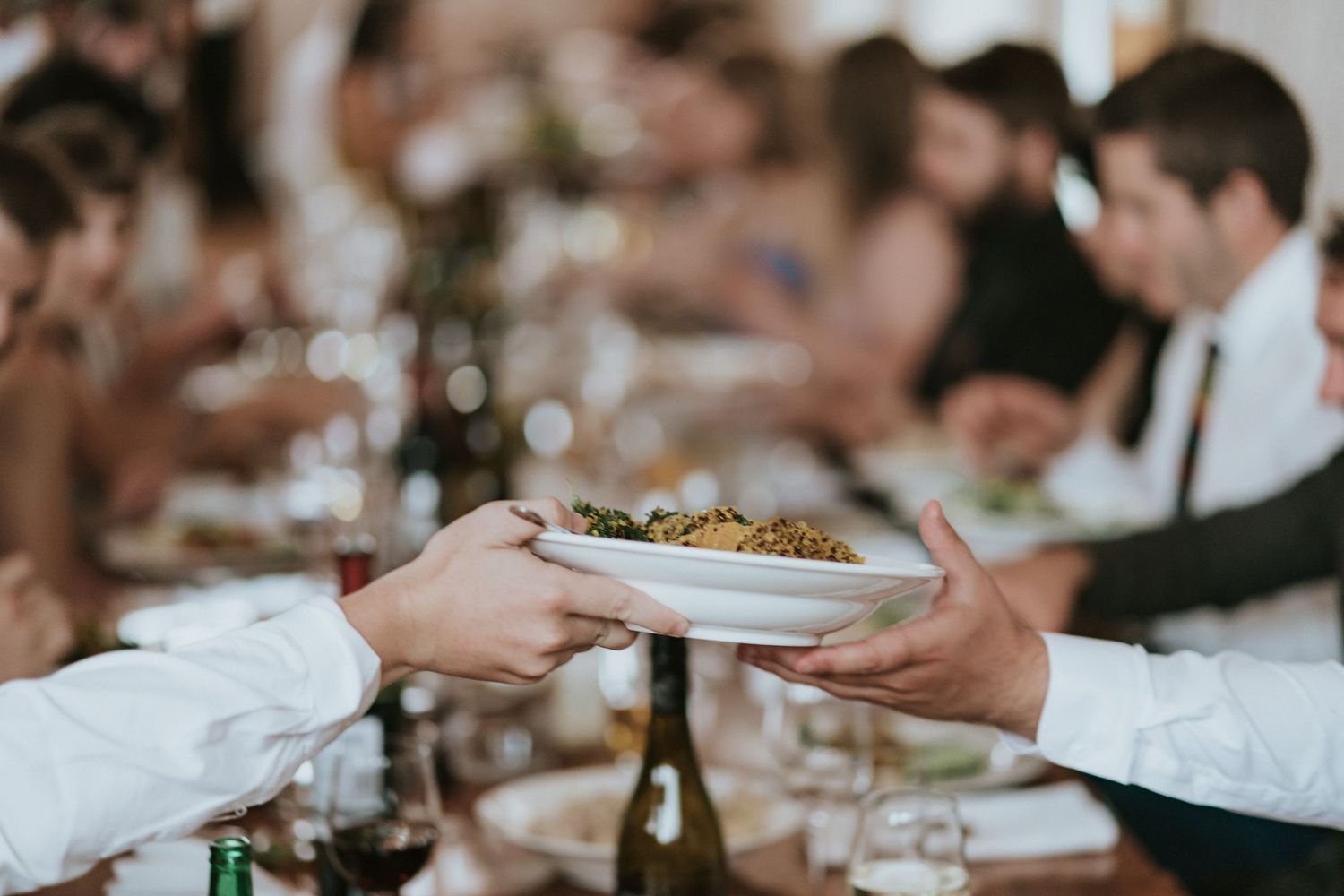 A server passes a plate of food to a guest at an elegant dinner party with wine glasses and table settings visible.