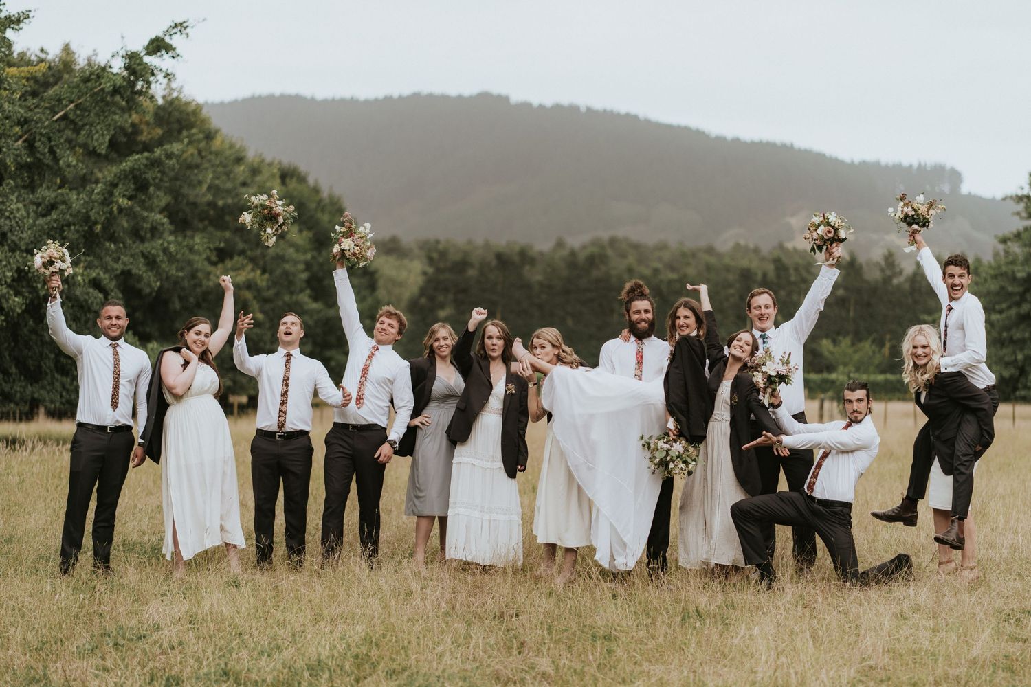 Wedding party celebrating outdoors with arms raised in joyful poses against scenic mountain backdrop in rustic field.