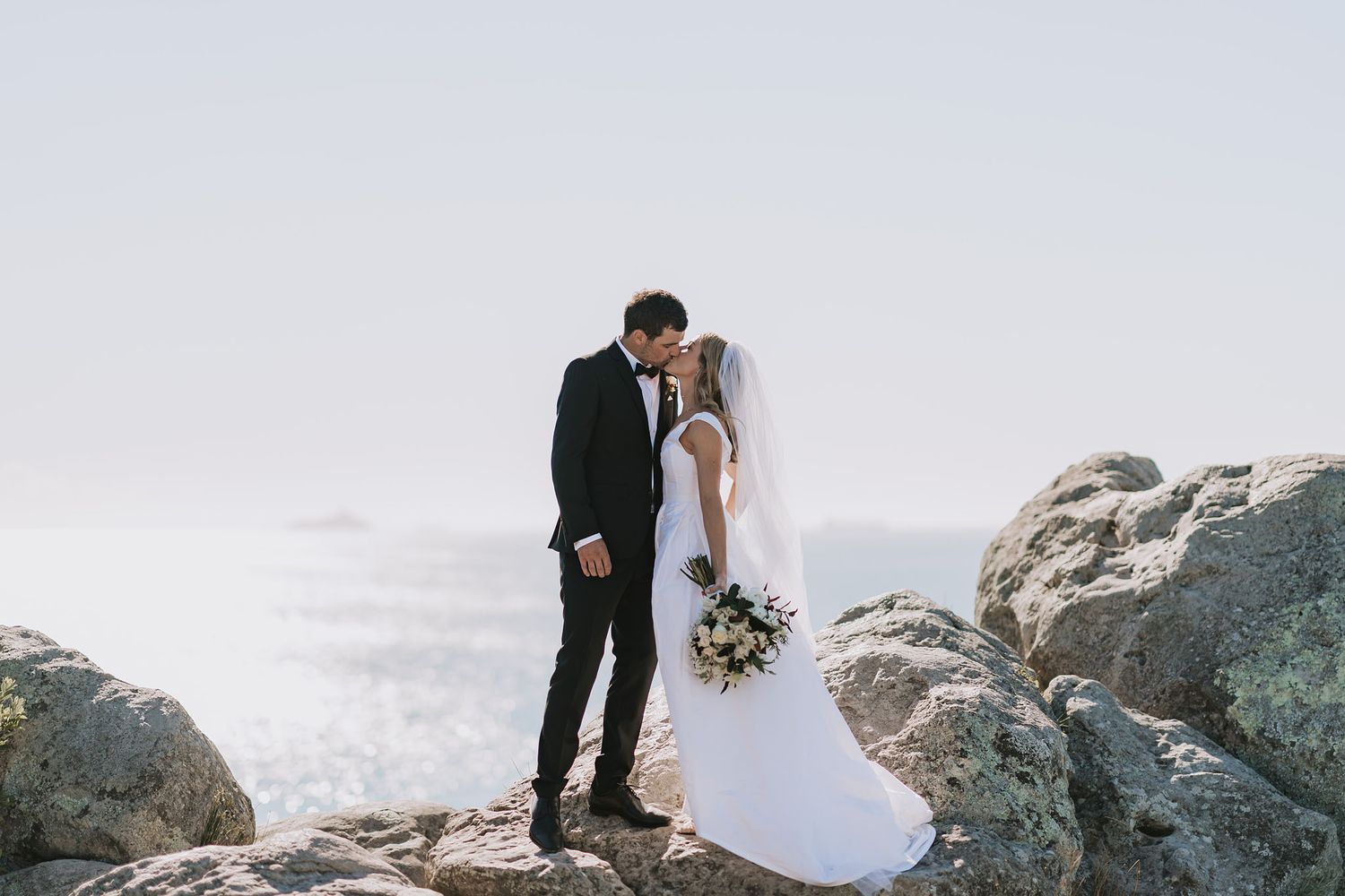 A romantic wedding couple standing on rocky cliffs overlooking a misty ocean view.