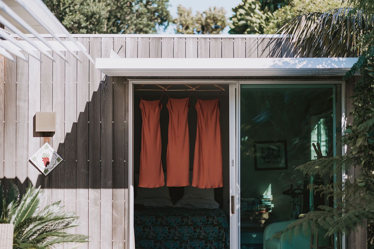 Three rust-colored dresses hang near a sliding glass door of a modern wood-paneled house with tropical plants outside.