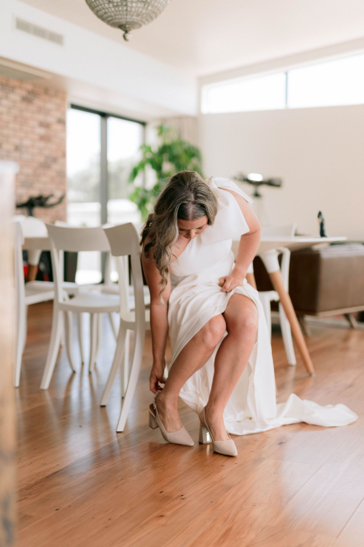 White-clad figure gracefully arranges dress on hardwood floor in bright modern interior with chandelier.
