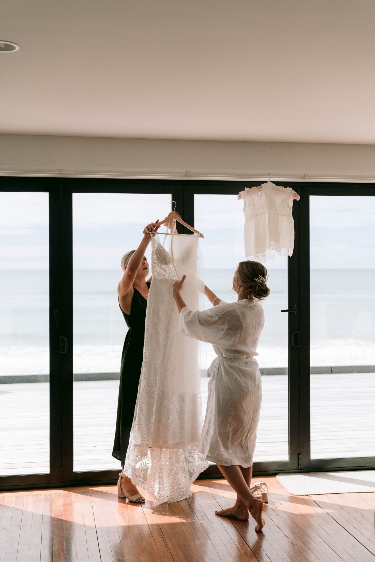 A person helps another get ready for a wedding by holding up a white wedding dress against a bright window backdrop.